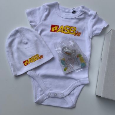 Babyset mit weißem Body, Mütze und Schnuller, alle mit ASBaby-Logo; daneben liegt eine weiße…