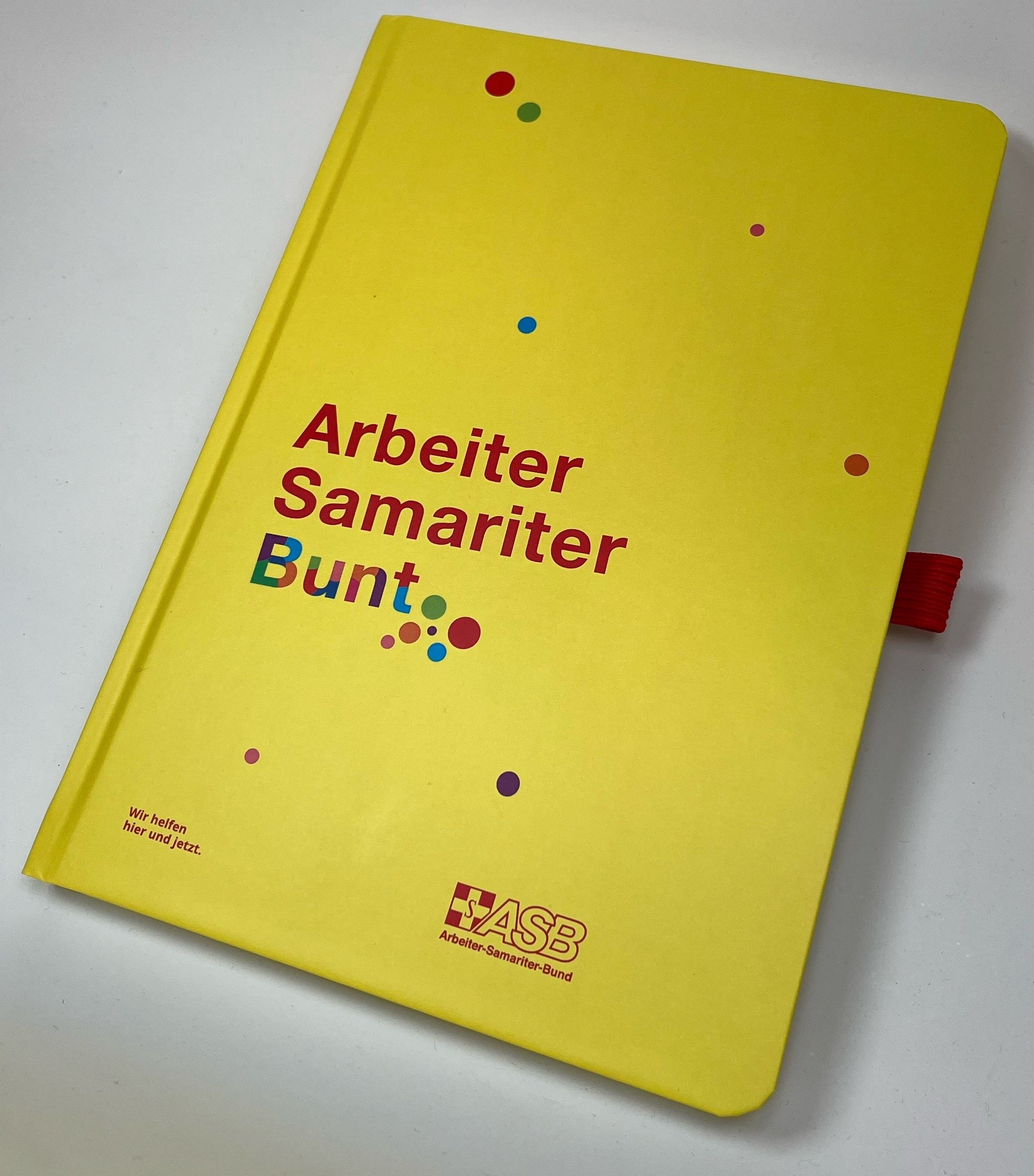 Notizbuch "ASB Bunt"  Gelbes Notizbuch mit der Aufschrift „Arbeiter Samariter Bunt“ und bunten Punkten, ASB-Logo…