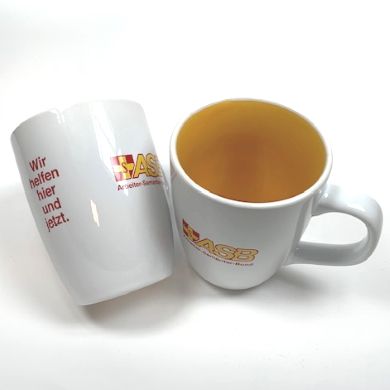 Zwei weiße Tassen mit gelber Innenseite, ASB-Logo und rotem Schriftzug „Wir helfen hier und…